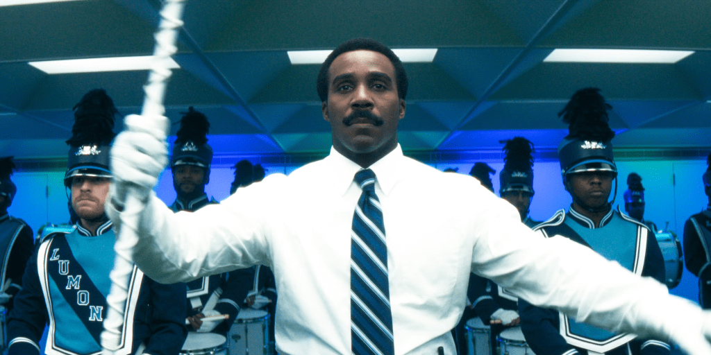 Mr. Milchick holds a baton and leads a band, the Choreography & Merriment Department